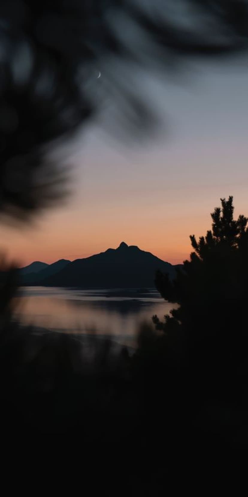 Dark mountain silhouettes and a crescent moon reflect on a still lake during a purple and orange sunrise.