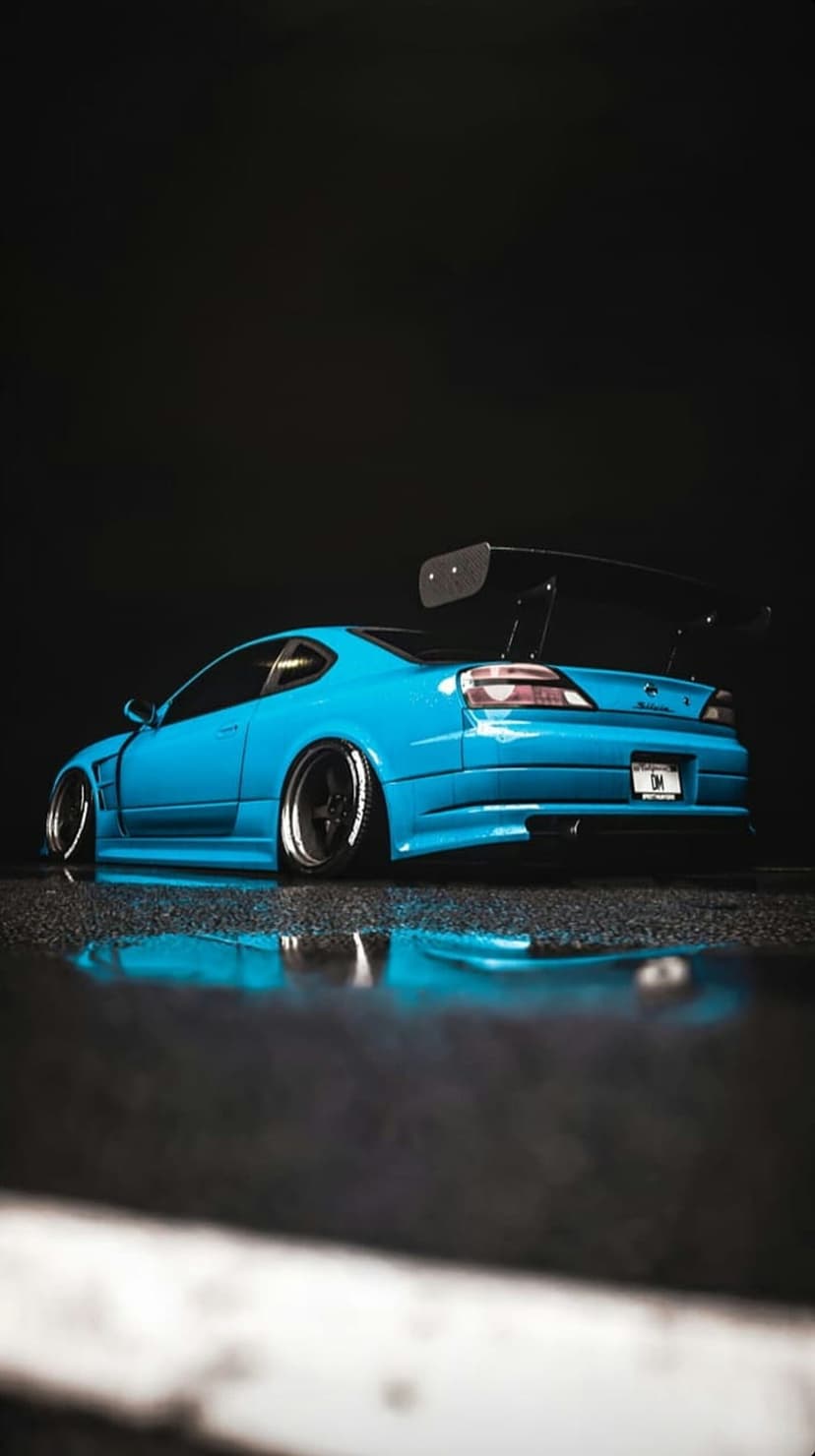 Blue Nissan Silvia S15 with a large spoiler and widebody kit sits on a wet dark road with vibrant reflections.