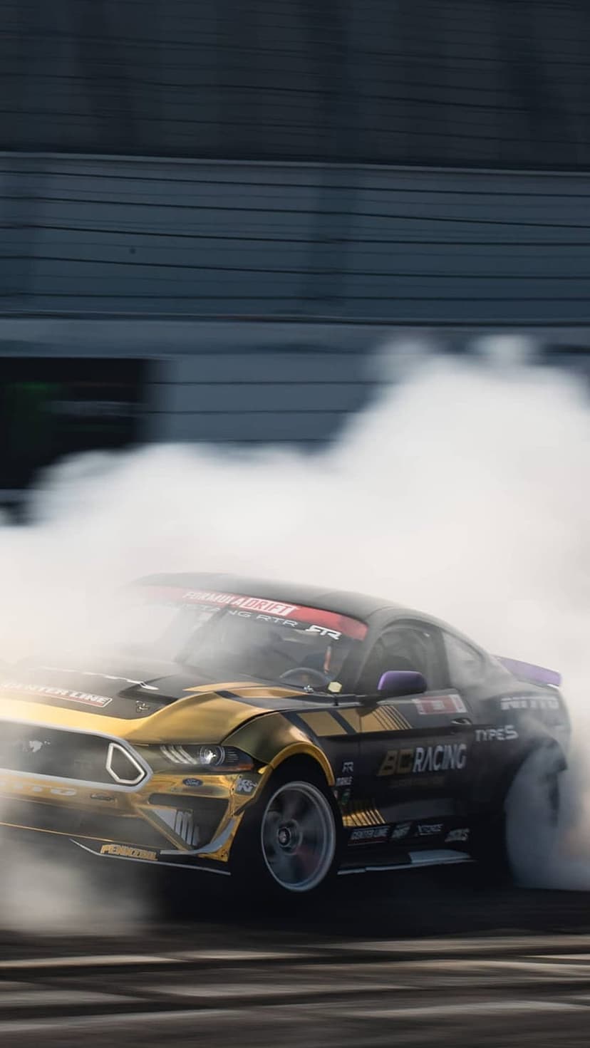 Gold and black Ford Mustang drifting on a race track with thick white tire smoke and motion-blurred background.