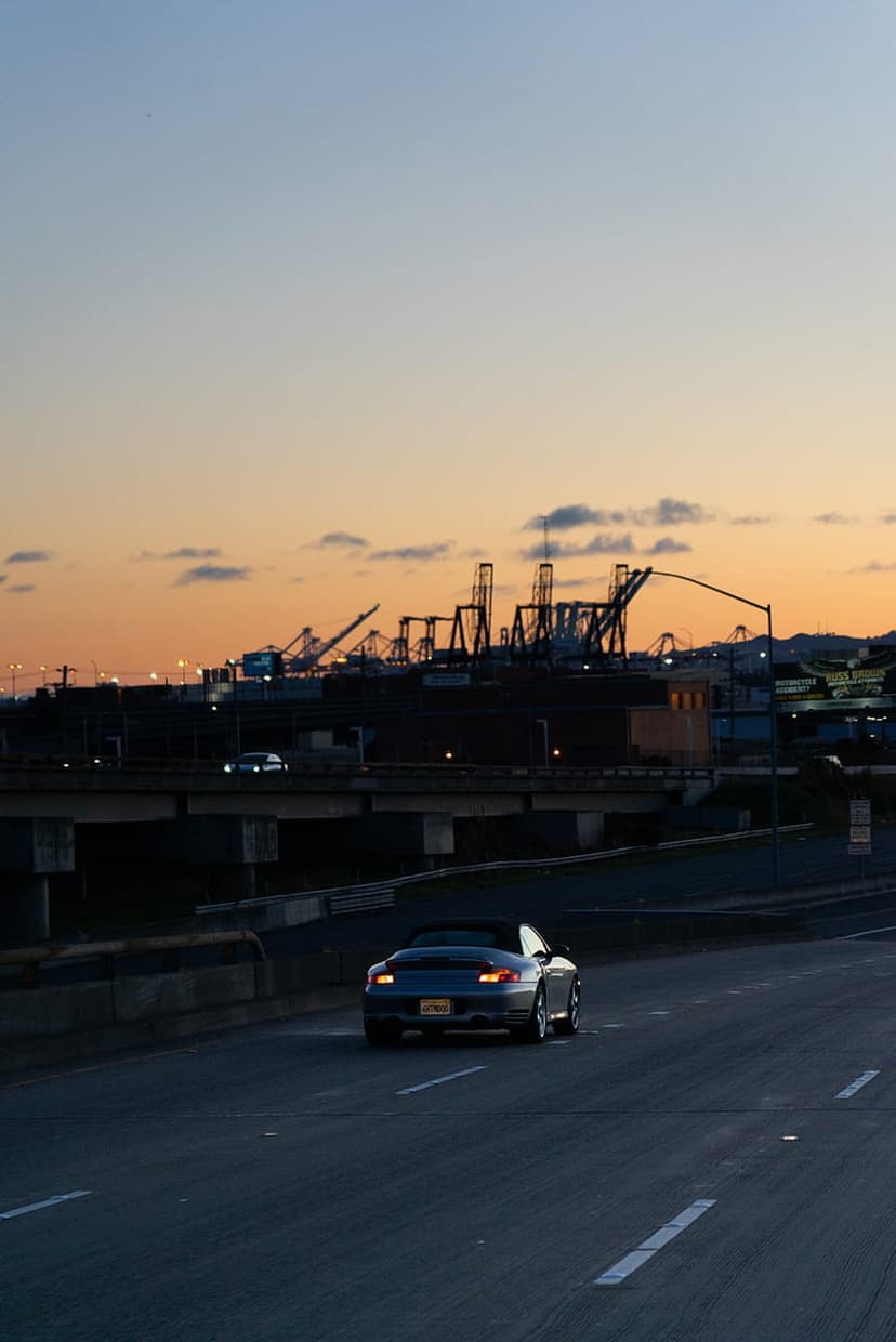 Convertible car driving on a highway at sunset with industrial port cranes silhouetted against a pink sky.