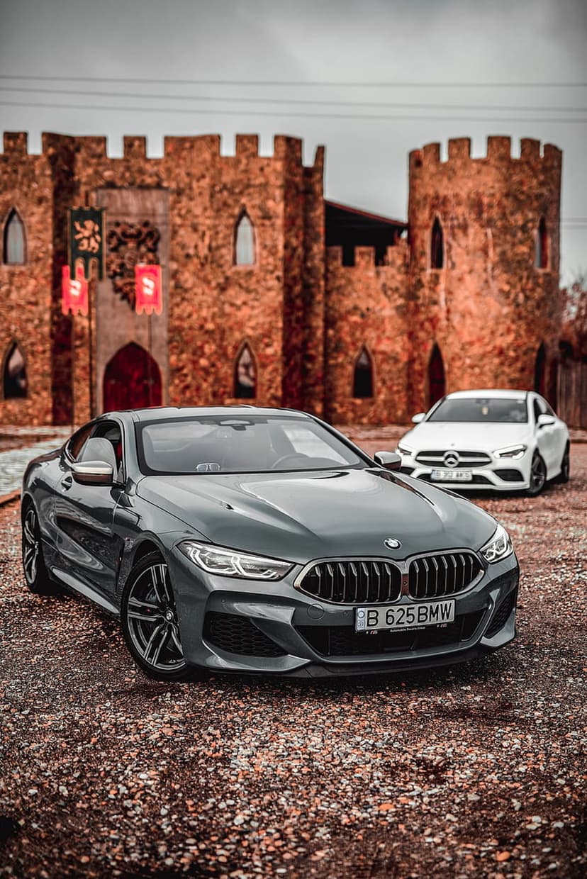 BMW and Mercedes-Benz cars parked at a stone castle with battlements under a cloudy sky during the autumn season.