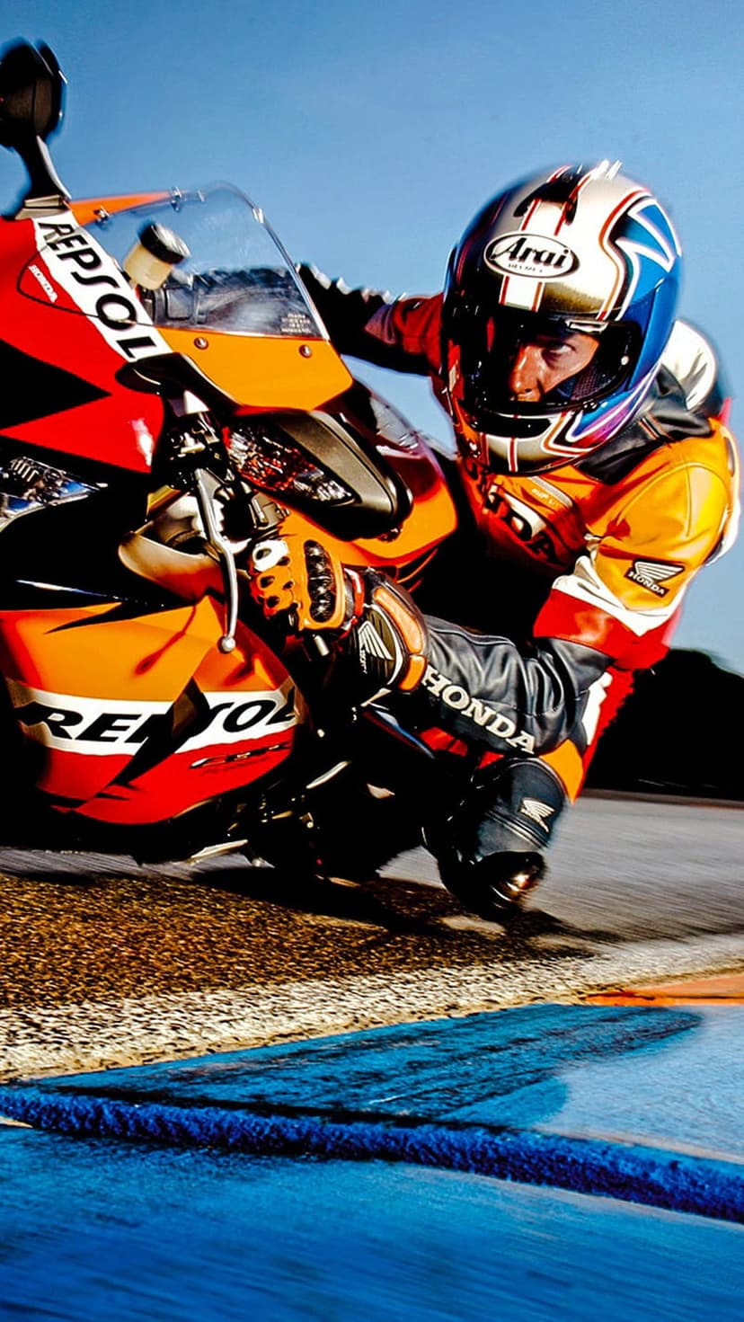 Motorcyclist in red and orange gear leans a racing bike low into a turn on an asphalt track under a blue sky.