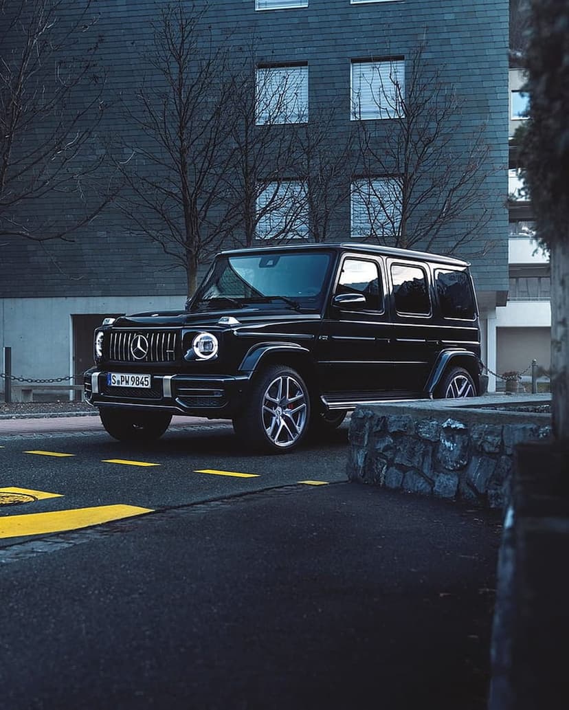 Black Mercedes-Benz G-Class SUV parked on a dark street with yellow road lines and leafless trees at twilight.