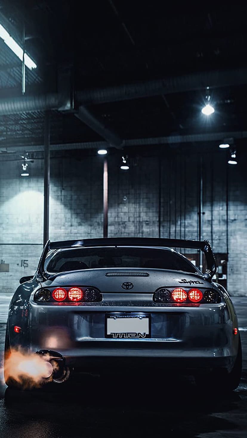 Toyota Supra rear view with orange exhaust flames and red brake lights in a dark industrial brick garage.