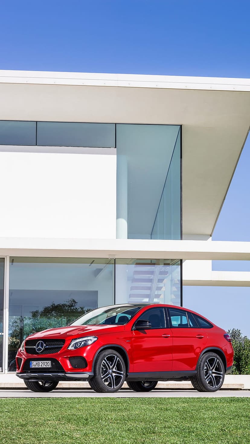Red Mercedes-Benz GLE Coupe parked on a green lawn in front of a modern white house with large glass windows.