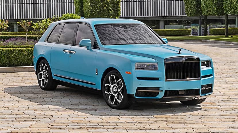 Light blue Rolls-Royce Cullinan SUV with black accents parked on a paved driveway with greenery in the background.