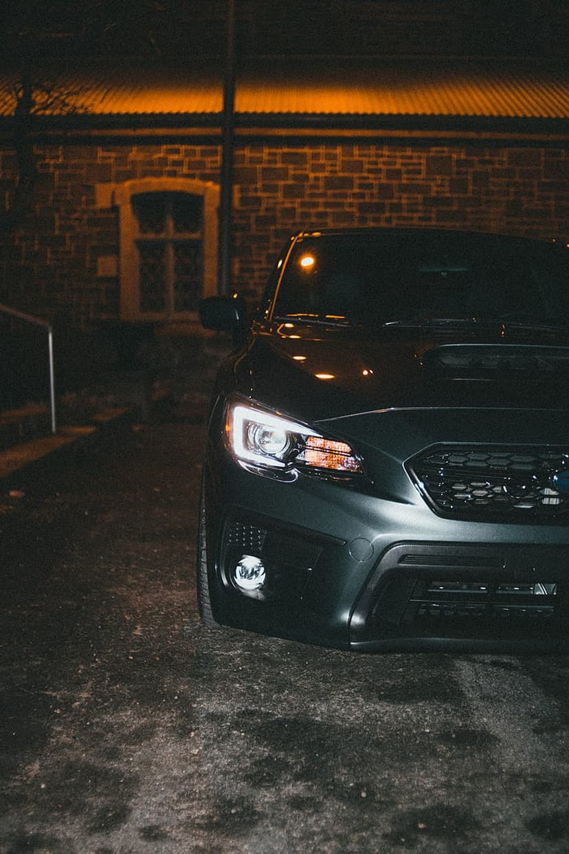 Dark Subaru WRX parked at night with headlights on against a brick building backdrop under warm orange light.