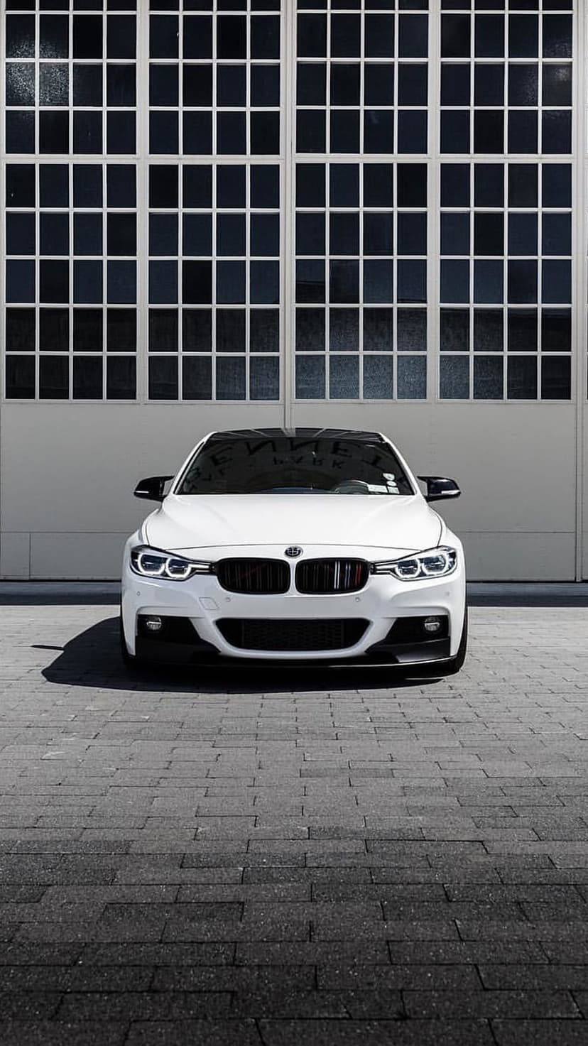 White BMW sedan with lit headlights parked on brick pavement in front of dark gridded industrial garage doors.