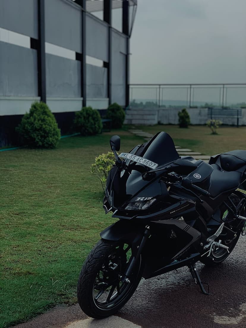 Black Yamaha R15 sports bike parked on vibrant green grass with a contemporary glass building in the background.