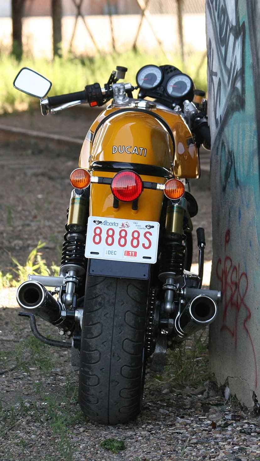 Yellow Ducati motorcycle rear view featuring a chrome exhaust and Alberta license plate against a graffiti wall.