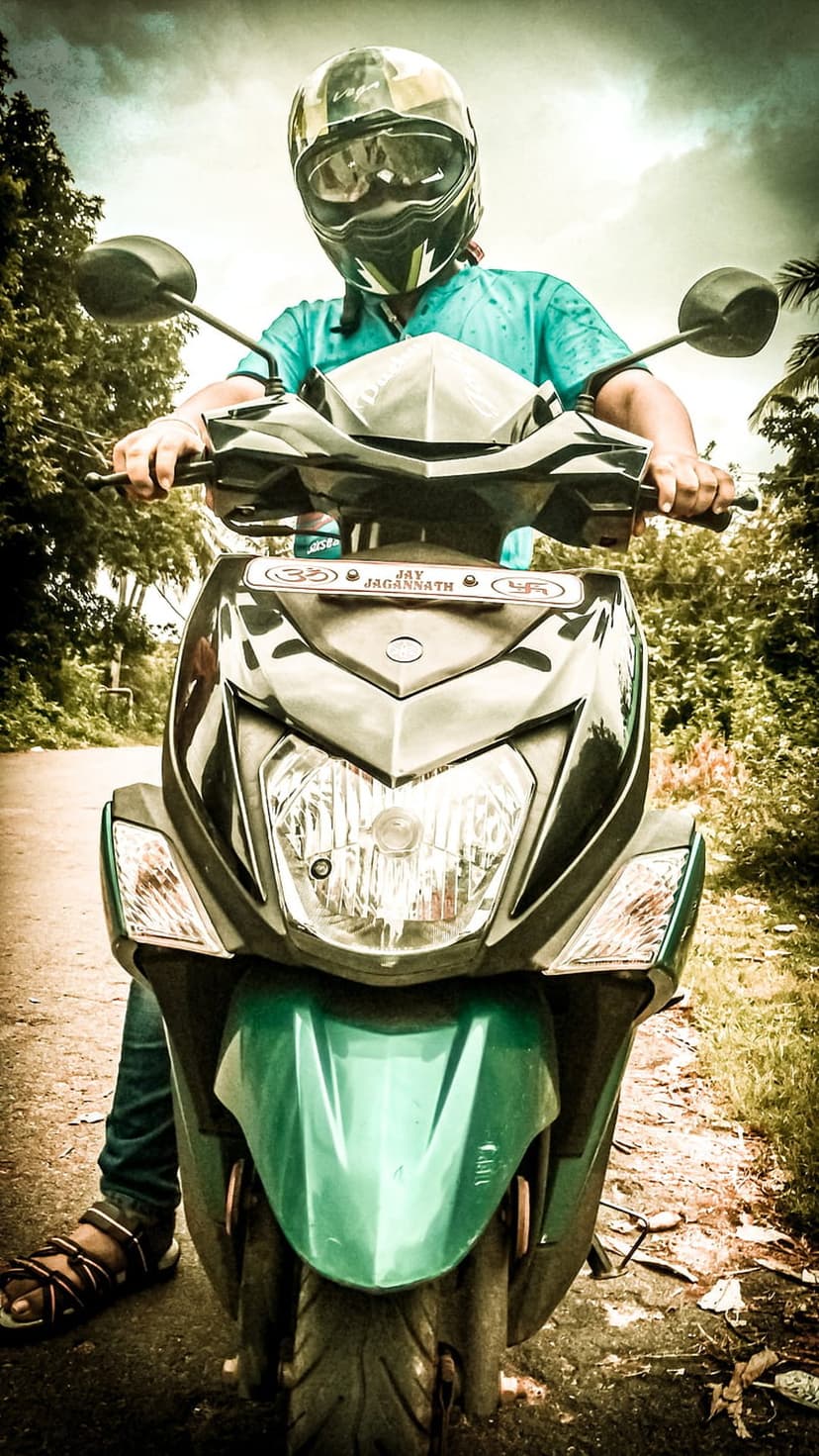 Person in a blue shirt and helmet riding a green scooter on a dirt road with trees under a cloudy sky.
