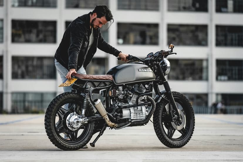 Man in a black jacket stands by a custom Honda motorcycle with a brown leather seat and grey tank in a city.