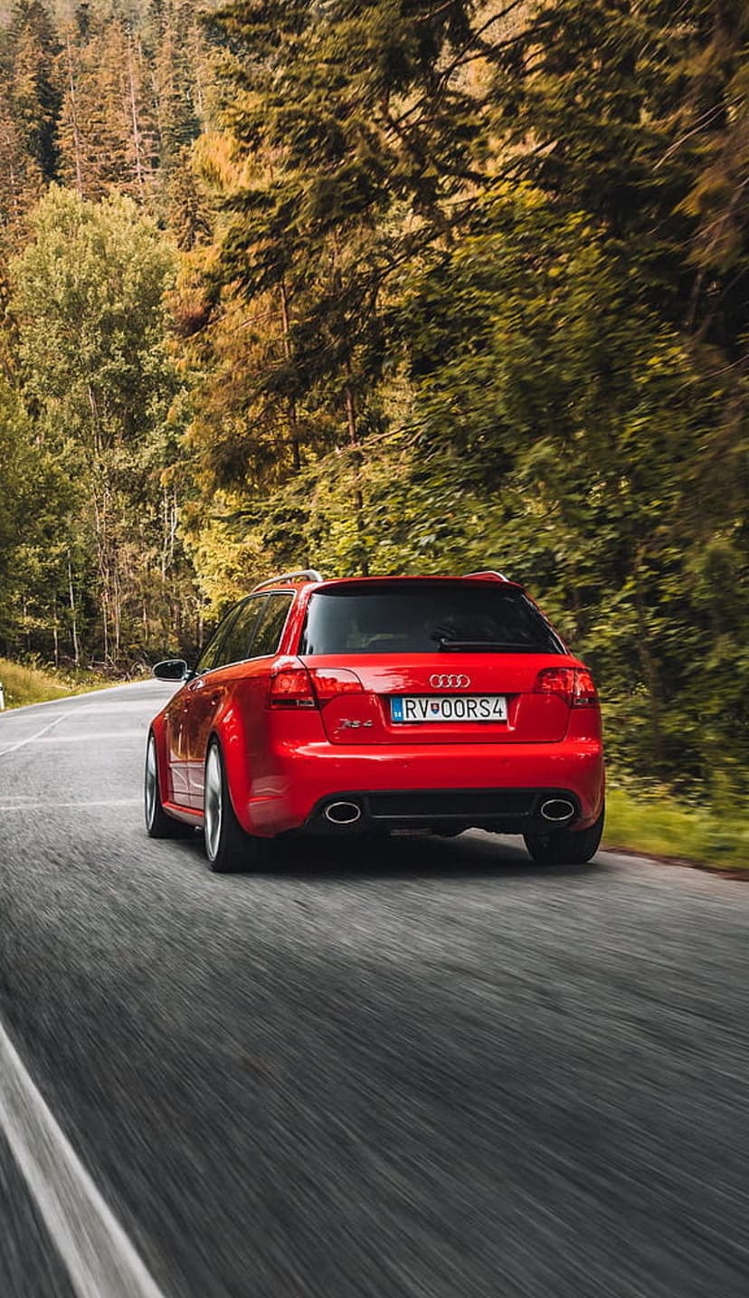 Red Audi RS4 Avant wagon drives along a winding road surrounded by orange and yellow autumn forest foliage.