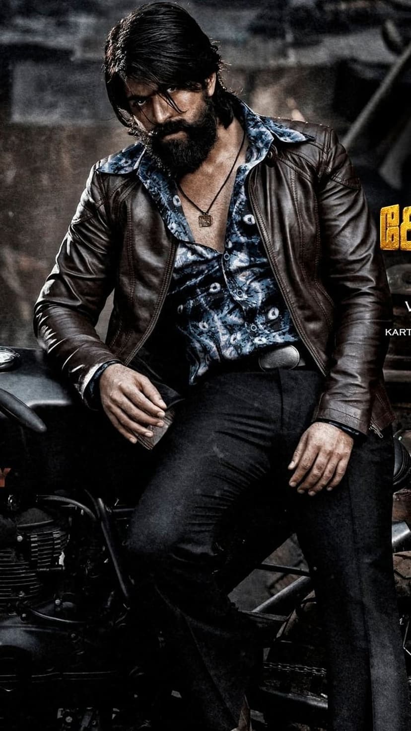 Yash as Rocky Bhai wears a brown leather jacket and patterned shirt with an intense gaze and full beard.