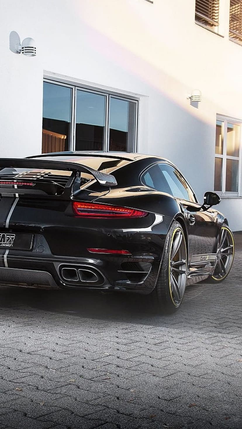 Black Porsche 911 Turbo S with a large rear wing and yellow-rimmed wheels parked on a stone driveway.