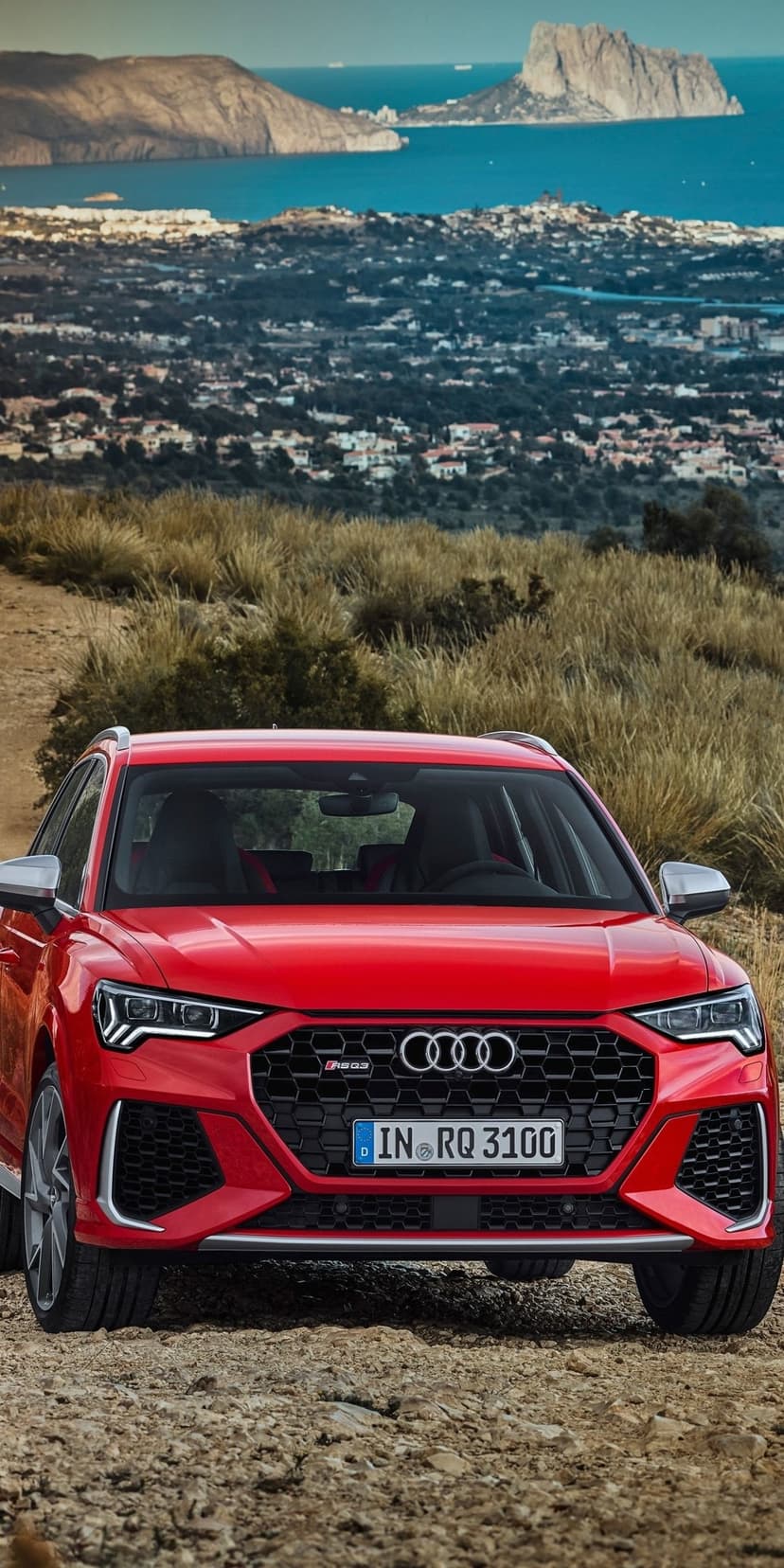Red Audi RS Q3 SUV on a dirt path with a coastal village and towering rock cliffs in the background.