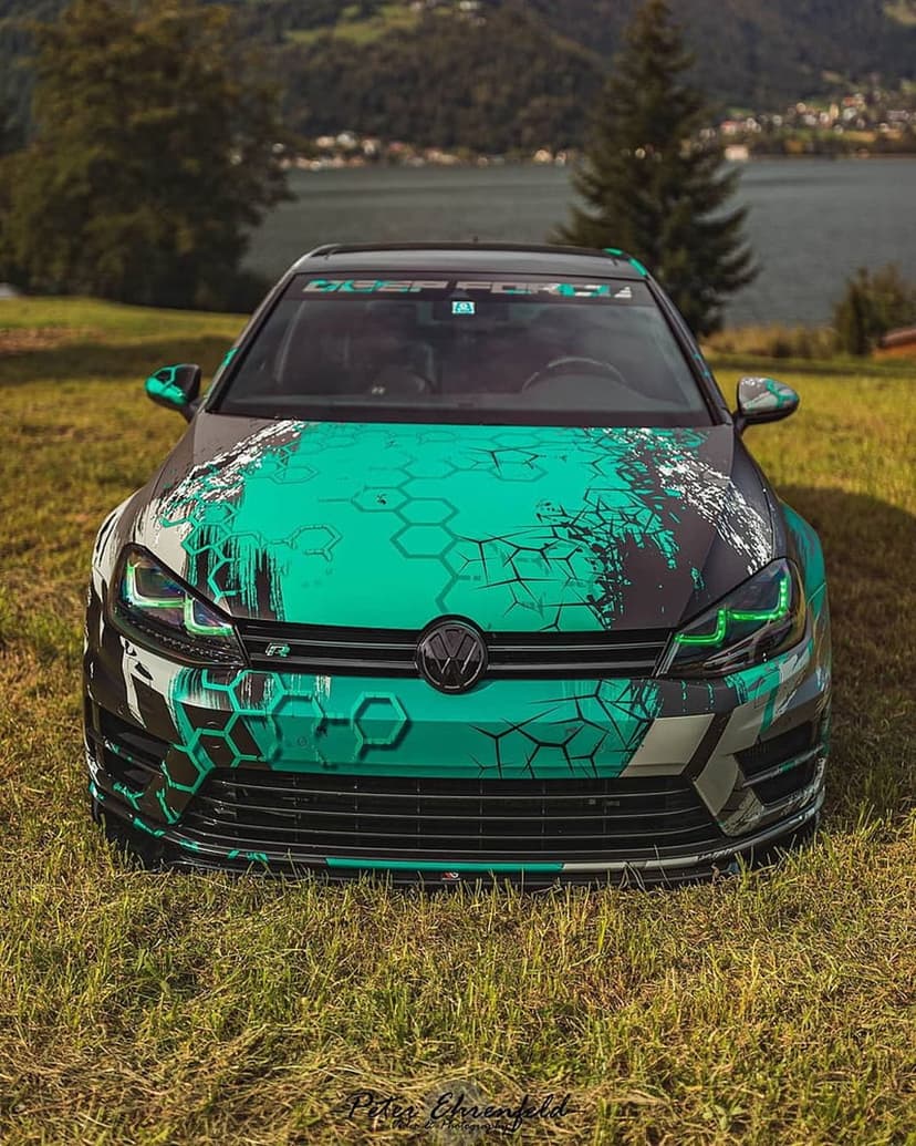 VW Golf R with a green and black hexagon wrap parked on a lush grassy hill overlooking a blurred scenic lake.