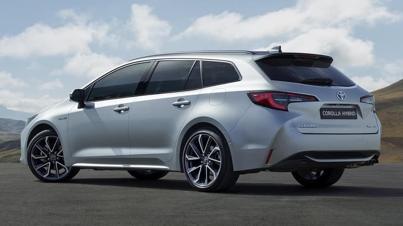 Silver Toyota Corolla Hybrid wagon parked on asphalt under a bright, cloudy blue sky in a wide scenic landscape.