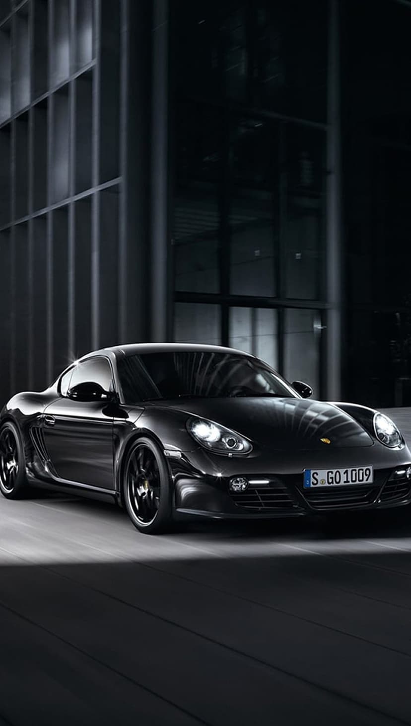 Black Porsche Cayman S driving past modern buildings at night with motion blur and glowing headlights.