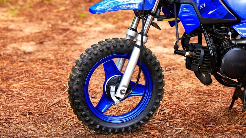 Close-up of a blue Yamaha dirt bike front wheel and silver forks on a ground covered in brown pine needles.