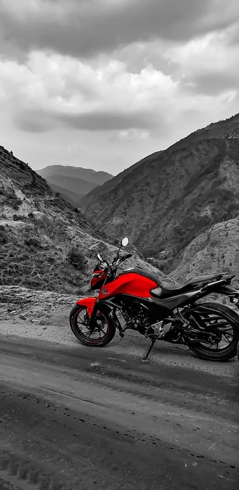 Red motorcycle parked on a rocky trail with dramatic grayscale mountain peaks and a stormy sky behind it.