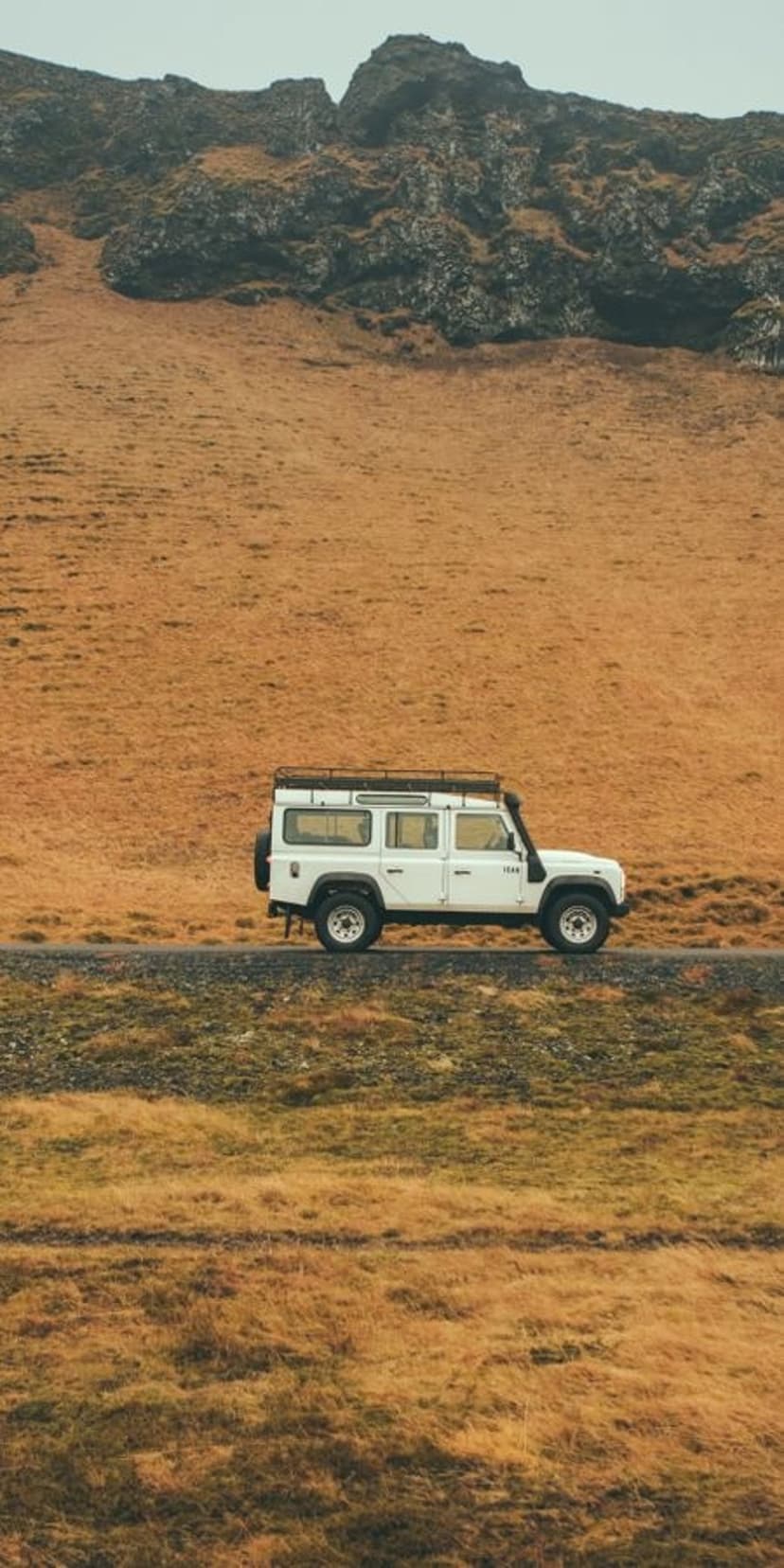 White Land Rover Defender 110 with a roof rack drives on a gravel road through Icelandic mountains and golden fields.