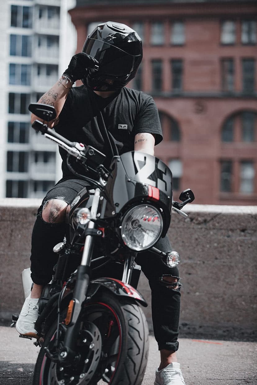 Biker in black gear and helmet sits on a custom motorcycle with red accents against an urban building backdrop.