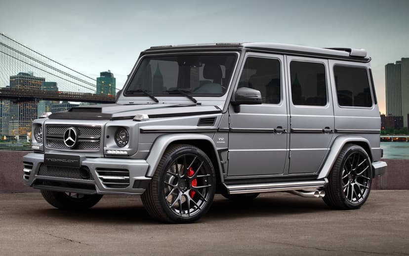 Silver tuned Mercedes G-Class SUV with carbon fiber accents and black rims parked near a bridge at dusk.
