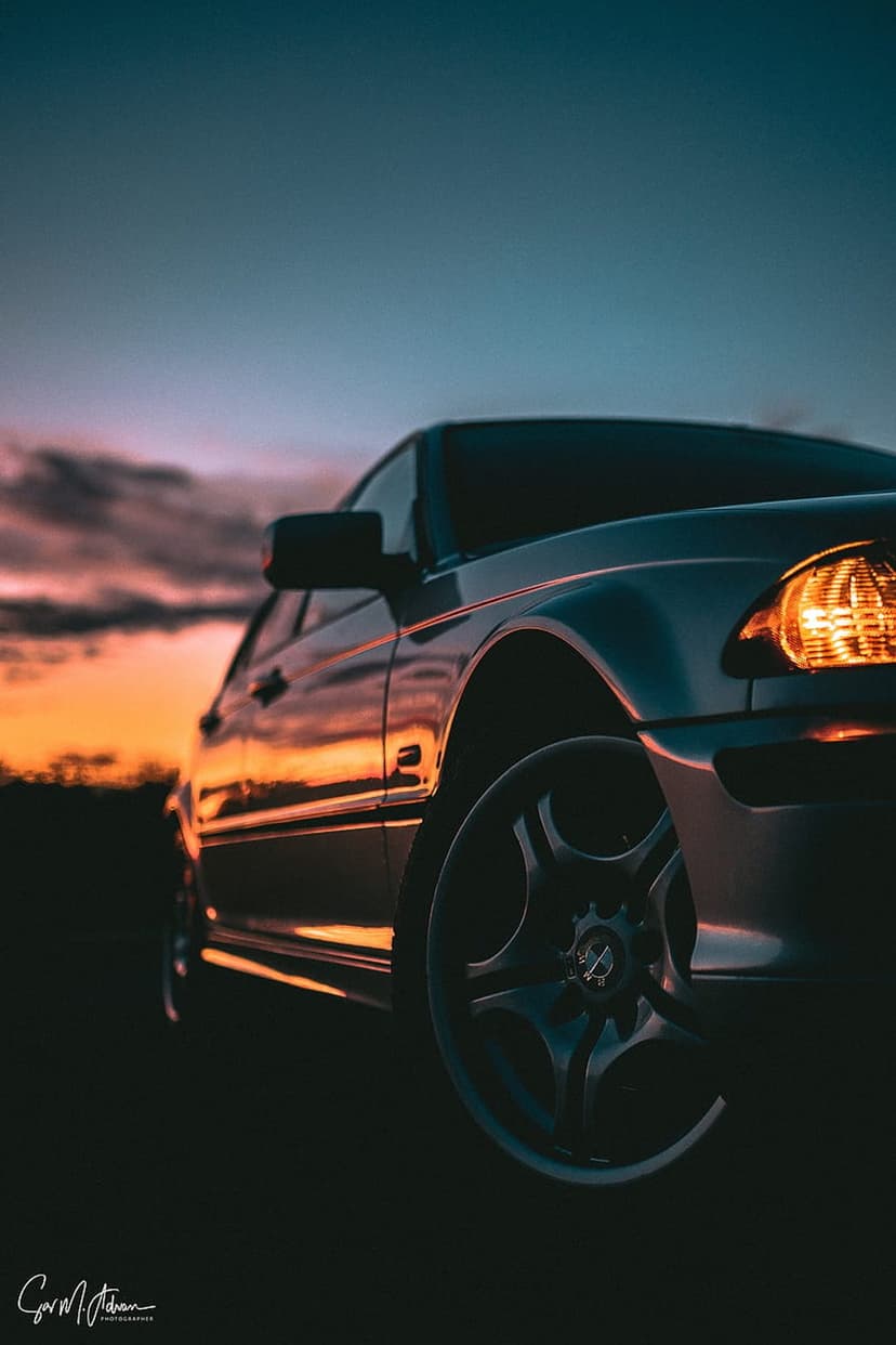 Low-angle car fender and alloy wheel reflecting orange sunset light against a dark twilight sky with headlights.