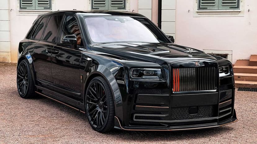 Black widebody Rolls-Royce Cullinan with orange grille accents parked on gravel against a light pink building.