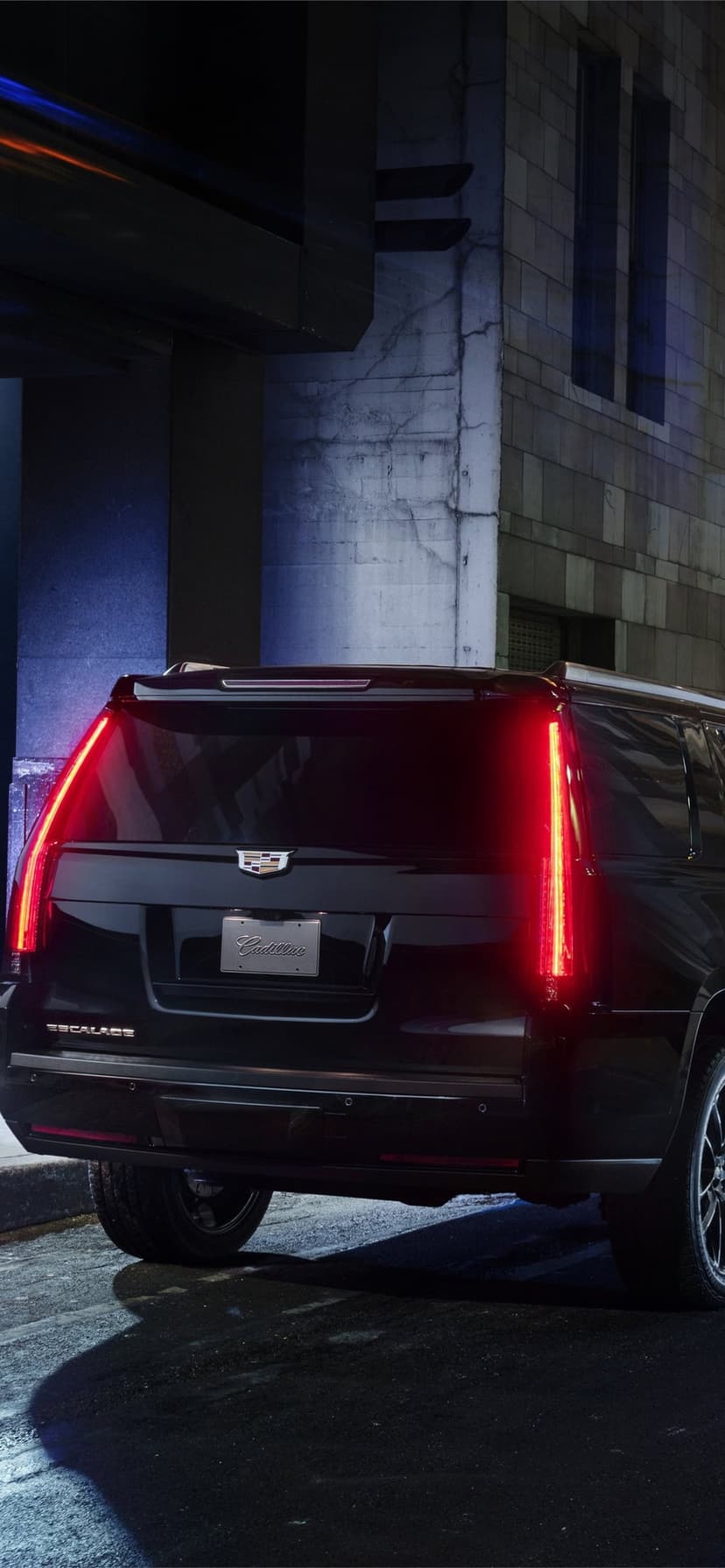 Rear view of a black Cadillac Escalade with glowing red vertical taillights in a dark city alley at night.