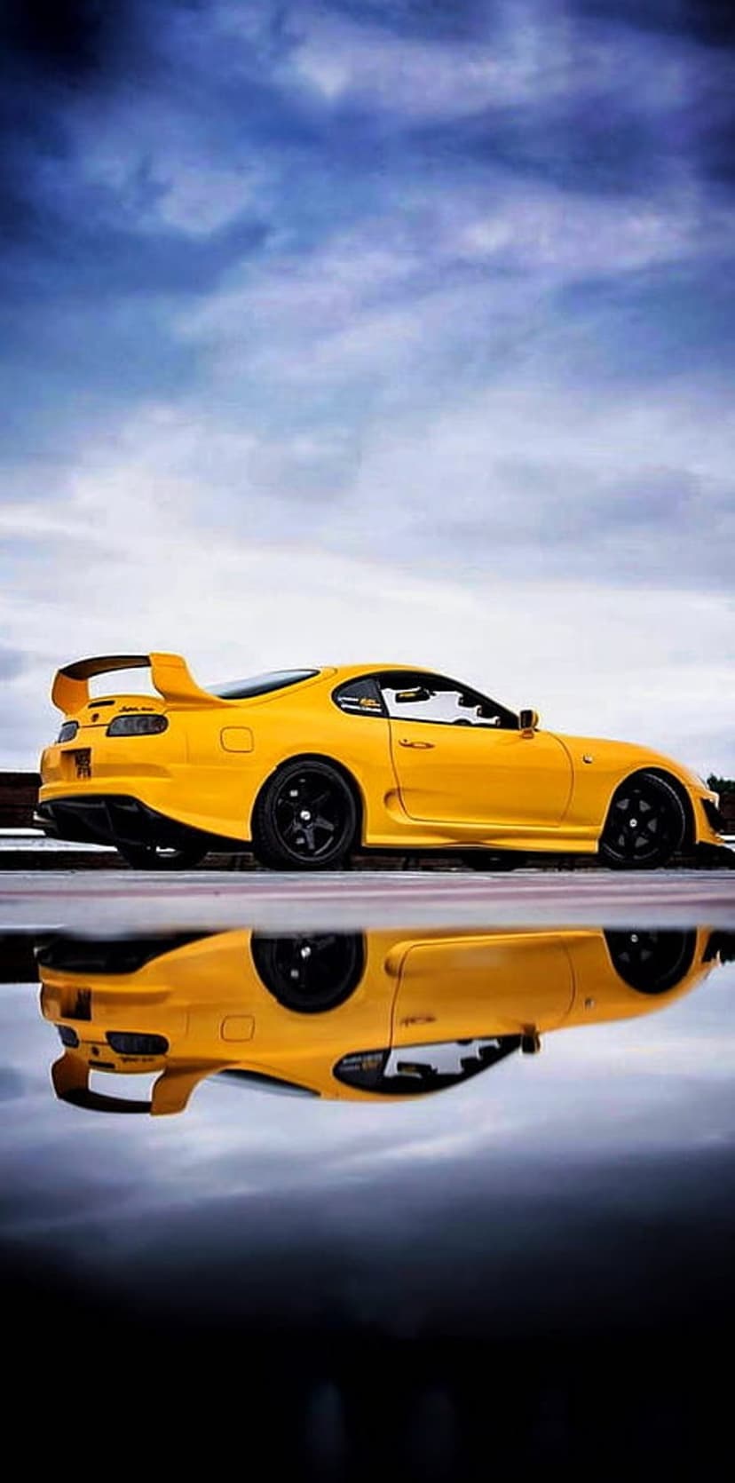 Yellow Toyota Supra MK4 with a large rear spoiler reflected on a wet asphalt surface under a gloomy sky.