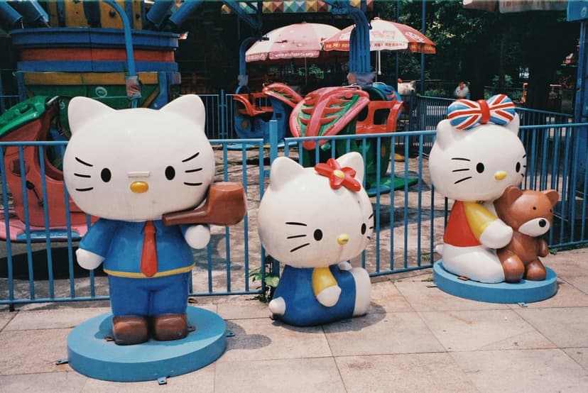 Hello Kitty statues in various outfits stand before amusement park rides and a blue fence on a sunny day.