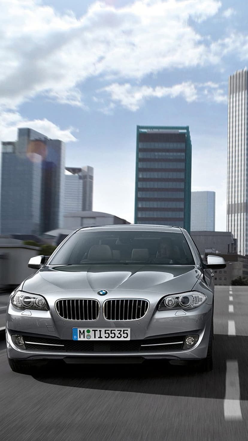 Silver BMW sedan with headlights on driving on an asphalt city road between tall glass skyscrapers and blue sky.