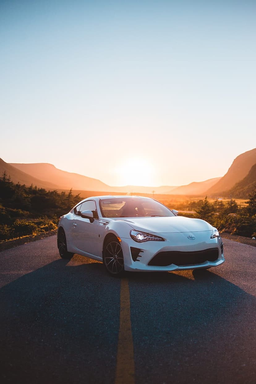 White sports car parked on a winding mountain road during a vibrant sunset with orange and purple sky.