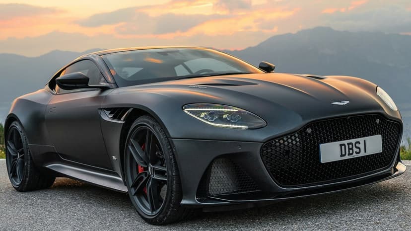 Matte black Aston Martin DBS Superleggera parked on a road with mountains and sunset sky in the background.