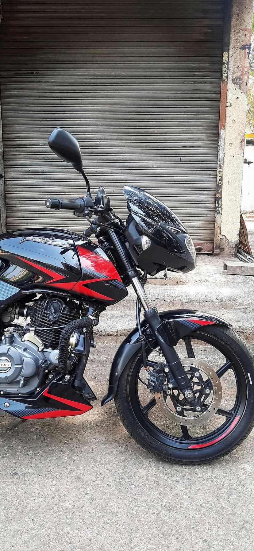 Black and red Pulsar motorcycle parked by a grey metal garage door showing the engine, disc brakes, and handlebars.