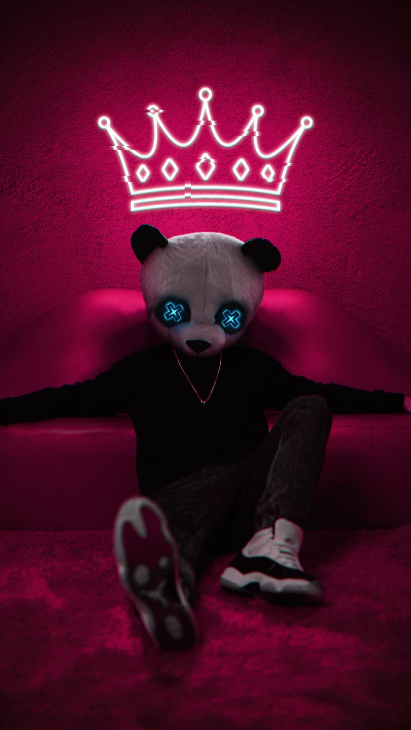 Panda mask with glowing blue eyes and a neon crown sits on a couch wearing black streetwear against pink.