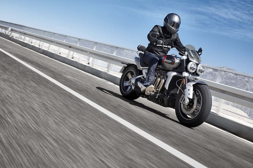 Silver Triumph Rocket 3 motorcycle speeds across a bridge under a blue sky with motion-blurred road surface.