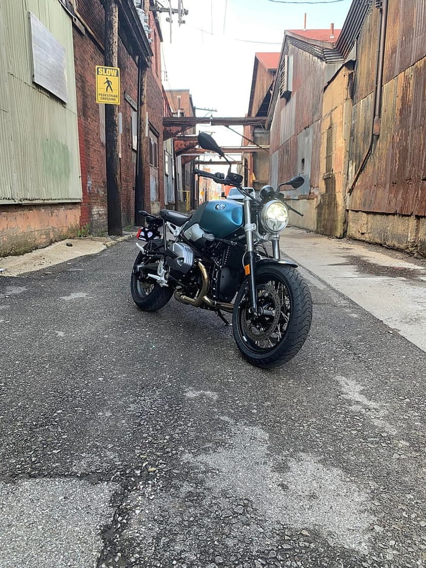 Teal BMW motorcycle with glowing headlight parked in a gritty alleyway near brick walls and a SLOW sign.
