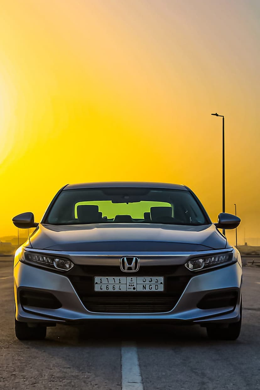 Silver Honda Accord sedan with glowing headlights faces forward against a golden orange sunset sky and street lamp.