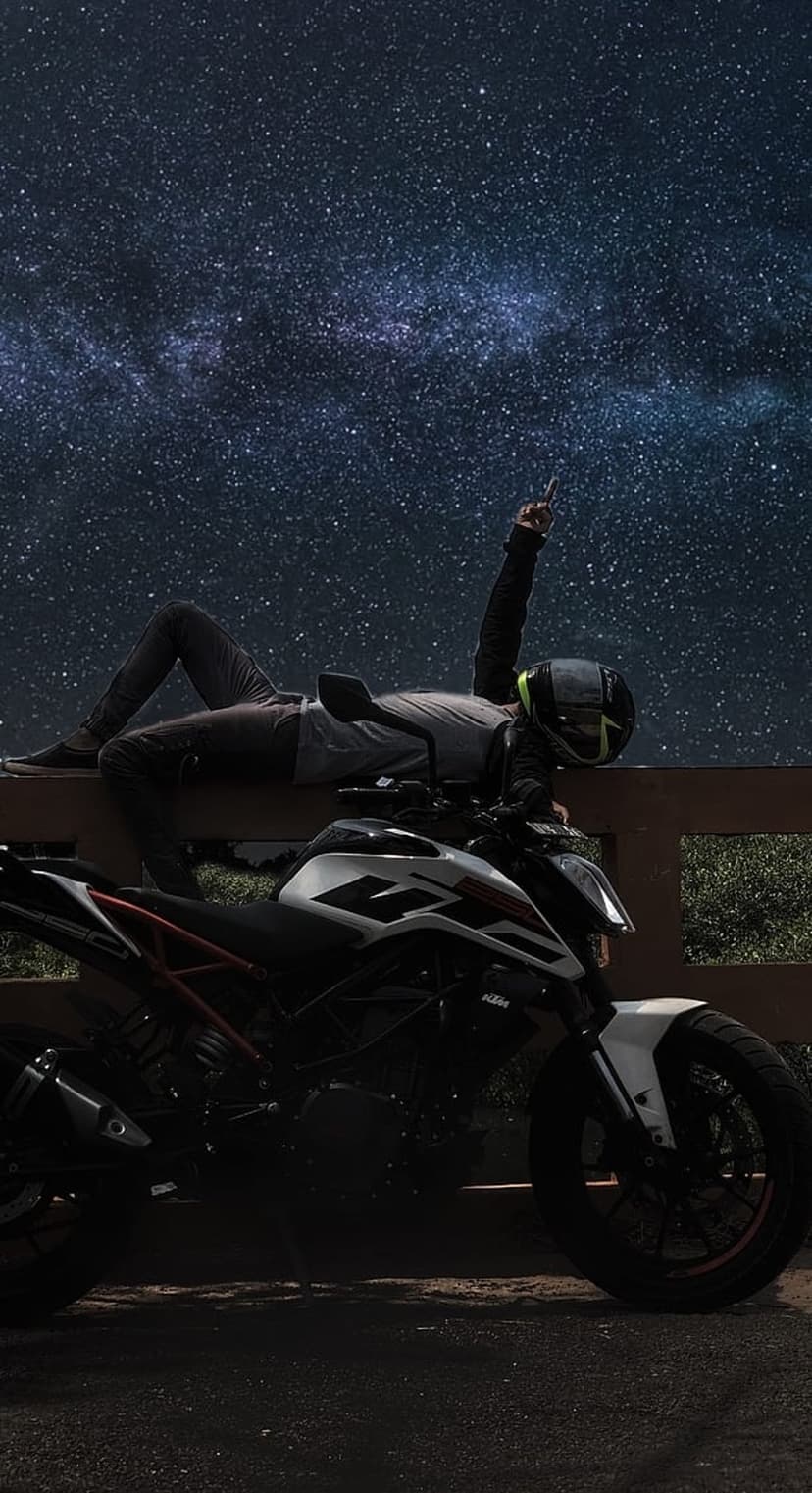 Motorcyclist in gear reclining on a wooden rail beneath a vibrant Milky Way galaxy and a parked bike at night.