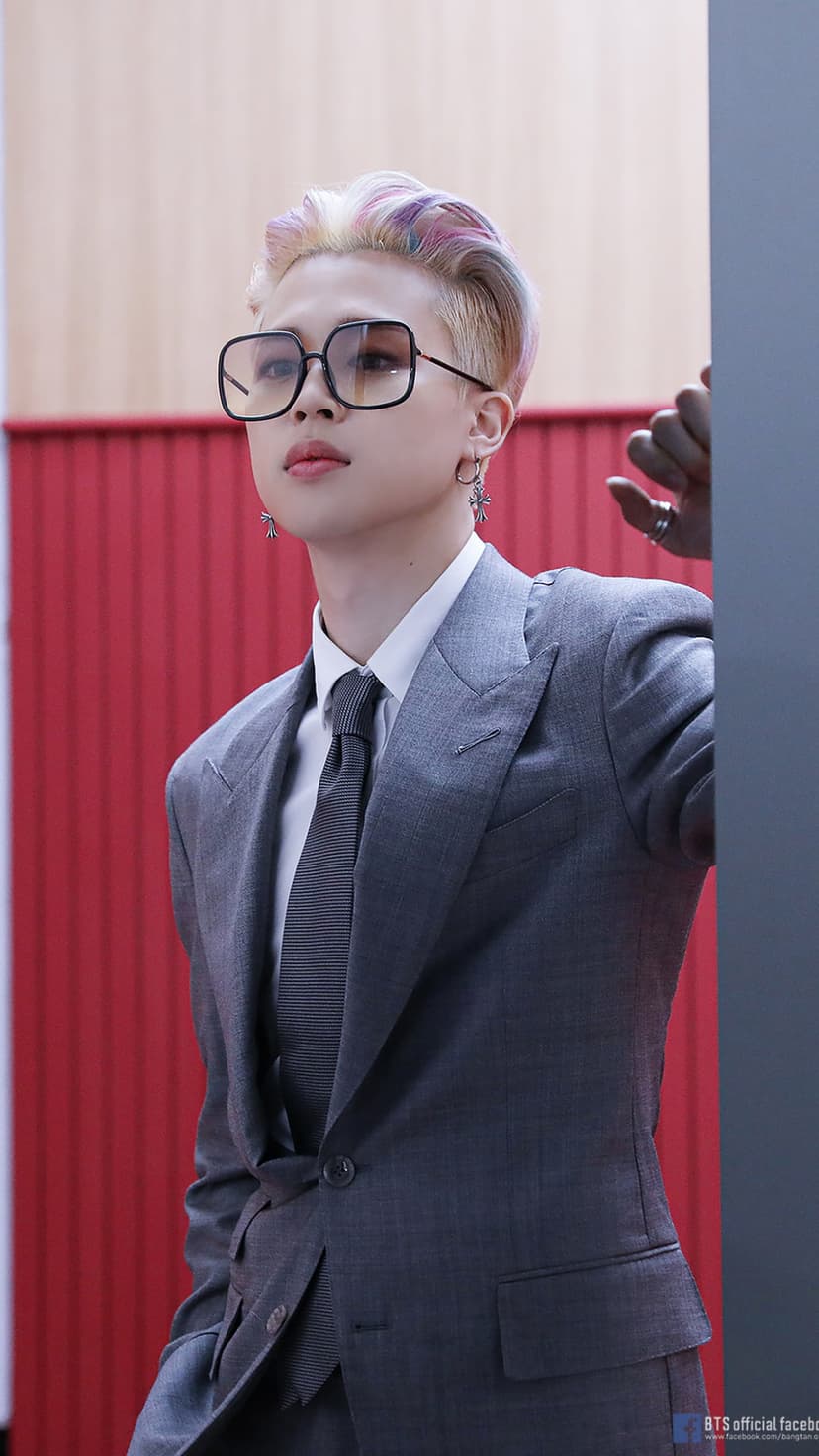 Jimin of BTS wearing a pinstripe suit and tinted square glasses against a red and grey textured background wall.