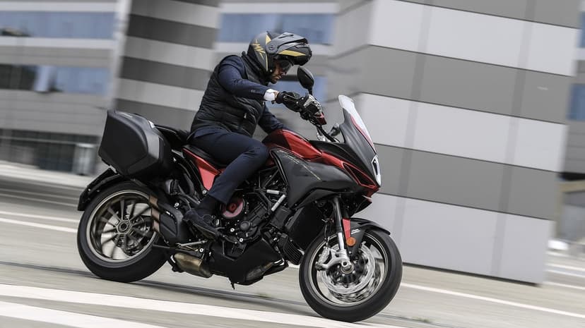 Red and black MV Agusta motorcycle with rider in motion past a modern building with geometric stripes.