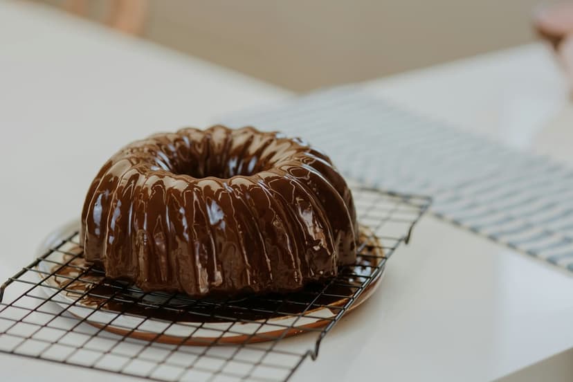 Chocolate Bundt cake with glossy dark glaze sits on a wire cooling rack under soft ambient lighting.