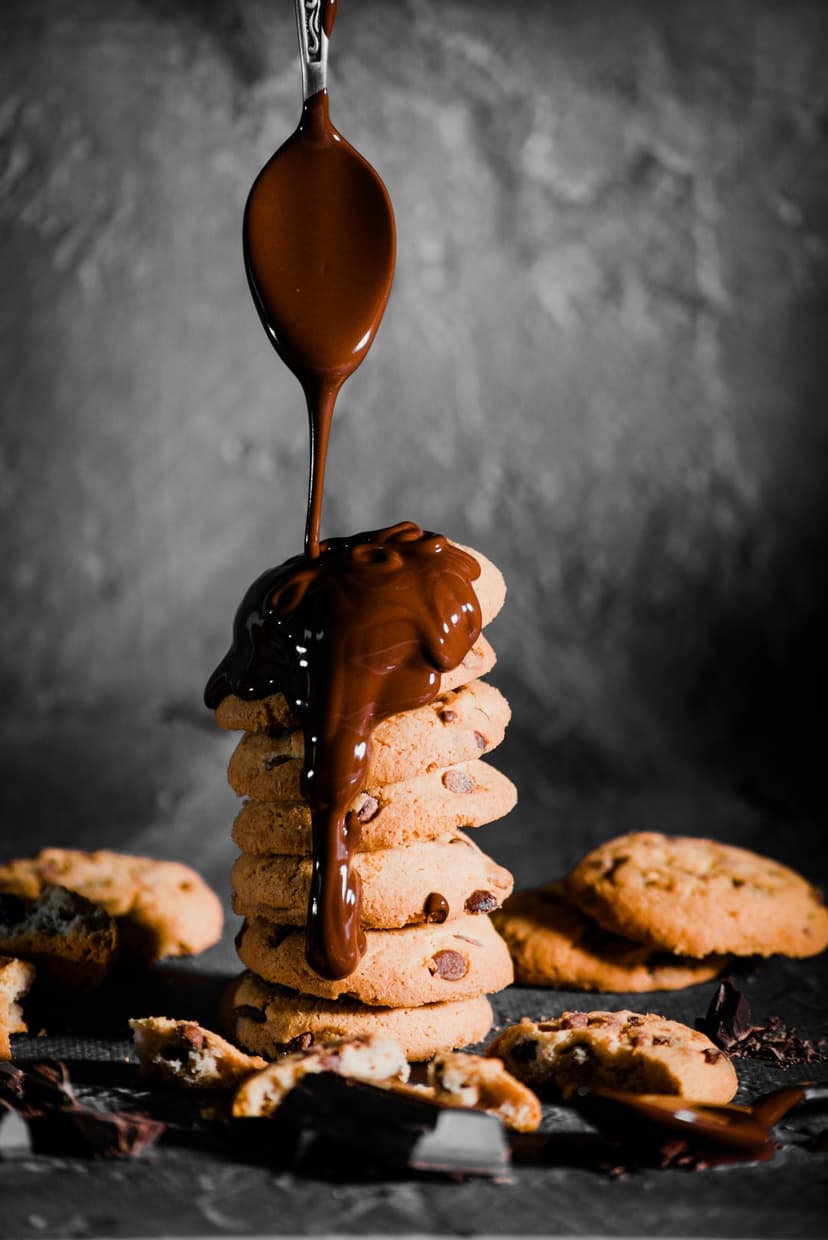 Molten chocolate sauce drizzling from a spoon onto a tall stack of golden brown chocolate chip cookies.