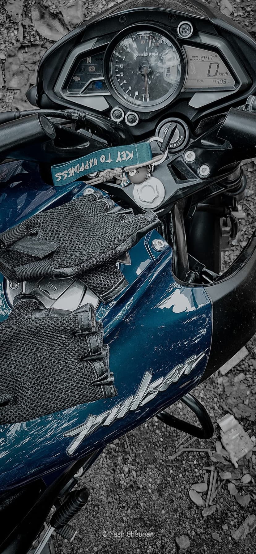 Blue motorcycle handlebars with black racing gloves, a digital speedometer, and a keychain on the fuel tank.