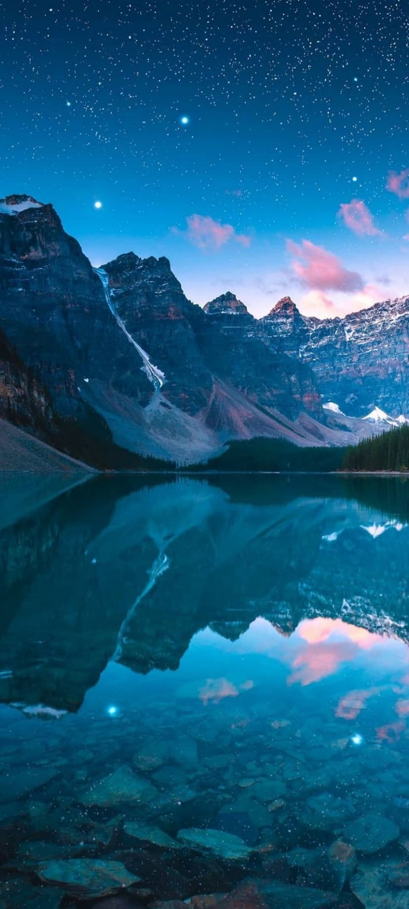 Snow-capped mountains reflect in a clear lake under a starry night sky with rocky terrain visible under water.