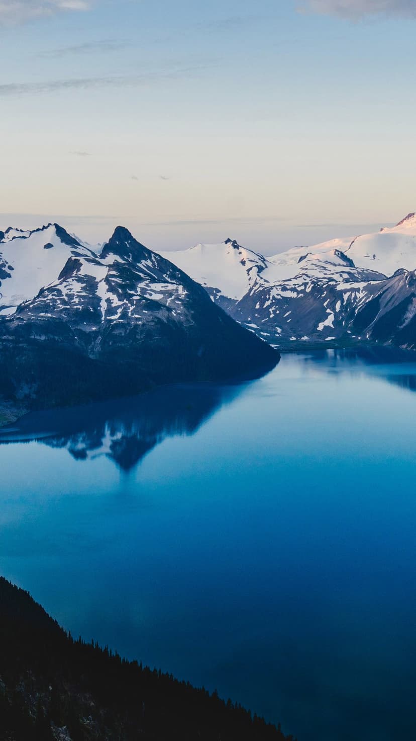 Snow-capped mountain peaks reflect perfectly in a still blue lake with a dark evergreen forest in the foreground.