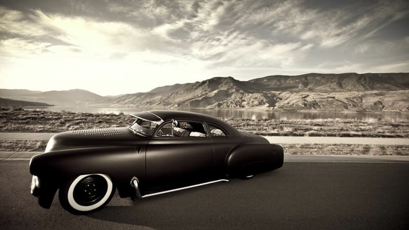 Low-profile matte black custom car with whitewall tires parked by a lake with mountains in the background.
